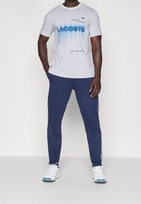 Grå T-shirt med blå Lacoste-texttryck, parat med navy sweatpants med sidostråkar och vita accenter. Sneakers med blå detaljer.