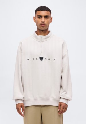 Jeune homme aux cheveux courts portant un pull Nike Golf beige clair avec une fermeture éclair sur le quart, debout devant un fond blanc uni.