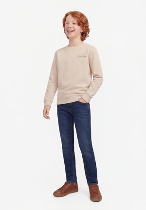 Red-haired boy smiling, wearing a beige sweatshirt, dark blue jeans, and brown sneakers, standing with one hand in pocket.