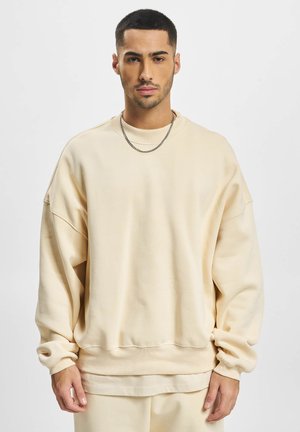 Beige oversized collegepaita, jossa on pyöreä kaula-aukko. Pehmeää puuvillakangasta, roikkuneet olkapäät ja resorit hihansuissa. Yhdistettynä hopeiseen ketjukaulaketjuun.