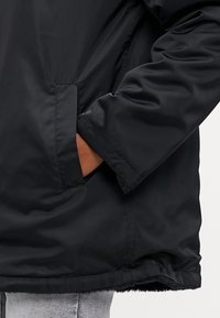 Ruka osoby v kapse černého zimního kabátu s viditelným lemem, v kombinaci se světle šedými kalhotami.