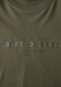 Olivově zelené tričko s vyraženým textem "Workshop Essentials Crafted Goods Unmatched Comfort Street One Men" uprostřed přední strany.