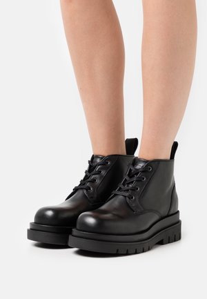 Botines de cuero negro hasta el tobillo con punta redonda, diseño de cordones, cuello acolchado y suela de goma texturizada con un agarre prominente, mostrados en piernas desnudas.