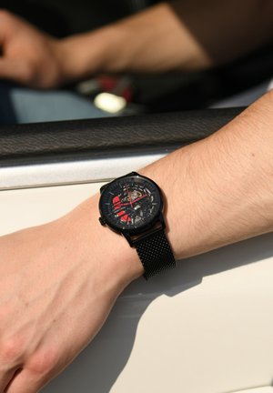 Montre noire avec un cadran transparent révélant des mécaniques complexes ; accents rouges et bracelet en maille noire, portée sur une main près de la fenêtre d'une voiture.