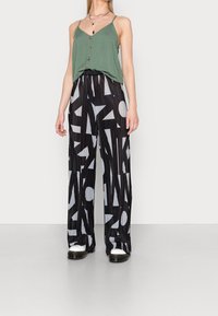 Top verde senza maniche con dettaglio dei bottoni, abbinato a pantaloni neri plissettati con un motivo geometrico in grigio e bianco.