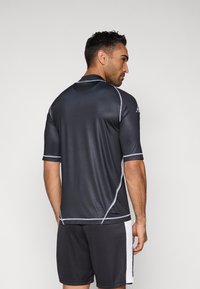 Temno siva atletska majica s kratkimi rokavi, s poudarki bele obrobe in ozekim krojem. Ujemajoče temne kratke hlače.