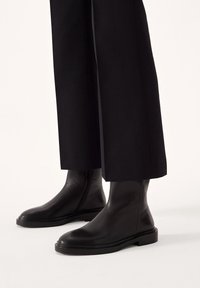 Czarne skórzane botki sięgające kostek o gładkiej powierzchni, strukturalnym kształcie, z bocznym zamkiem błyskawicznym oraz płaską gumową podeszwą, zestawione z szerokimi czarnymi spodniami.