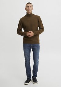 Pull en tricot marron à col roulé avec des poignets et un ourlet côtelés, associé à un jean slim bleu et des baskets noires sur fond blanc uni.