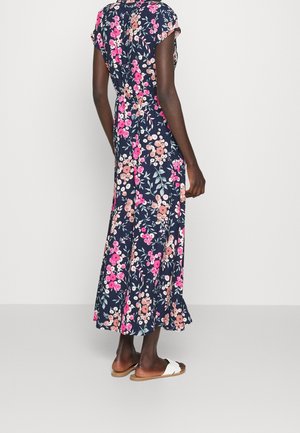 Kobieta stojąca tyłem, ubrana w granatową sukienkę midi w kwiaty oraz białe płaskie sandały na jednolitym jasnym tle.