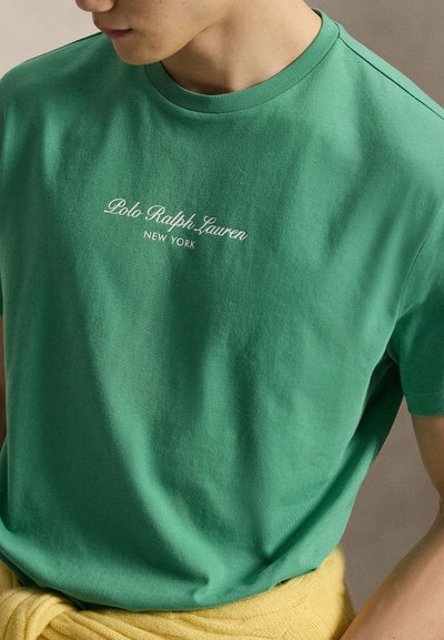 Άτομο που φοράει πράσινο μπλουζάκι Polo Ralph Lauren New York με ένα κίτρινο πλεκτό ρούχο δεμένο γύρω από τη μέση.