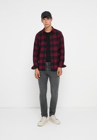 Camisa de cuadros rojo y negro sobre una camiseta negra, jeans grises y zapatillas blancas. La camisa tiene botones y las mangas arremangadas. Se lleva una gorra negra.