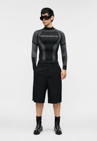 Μαύρο μακρυμάνικο rash guard με επενδυμένες υφές και λογότυπο· συνδυάζεται με μαύρο σορτς μέχρι το γόνατο και γυαλιστερά μαύρα μποτάκια αστραγάλου.