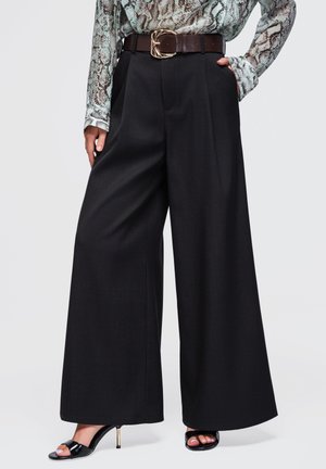 Femme portant un pantalon noir à jambes larges, un chemisier à motif peau de serpent, une ceinture marron avec une boucle dorée et des talons ouverts noirs.