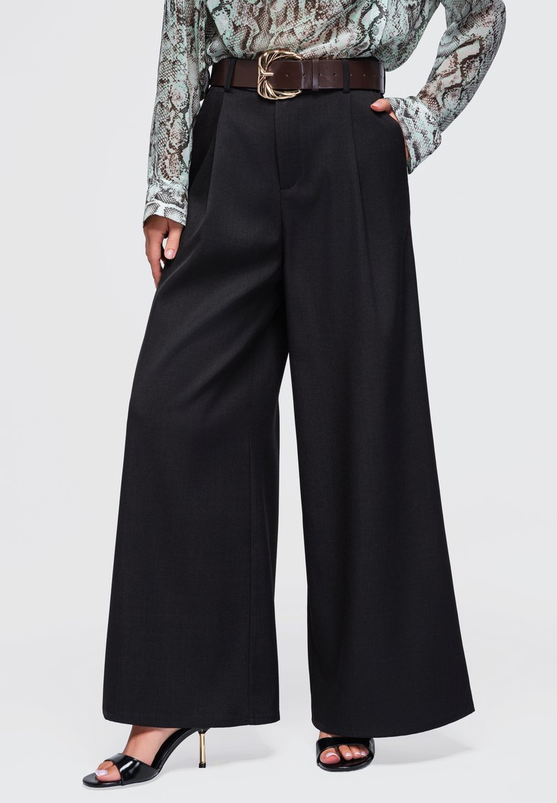 Femme portant un pantalon noir à jambes larges, un chemisier à motif peau de serpent, une ceinture marron avec une boucle dorée et des talons ouverts noirs.