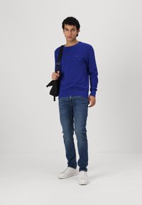 Pull bleu à col rond en tissu doux, associé à un jean bleu clair et des baskets blanches. Le pull présente un petit logo sur la poitrine.