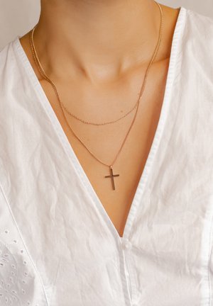 Gold-tone layered necklaces featuring a thin chain with a small cross pendant. Worn over a white, textured, v-neck top.