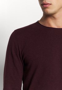 En ung person klädd i en långärmad, mörkt vinröd tröja med rund halsringning, visad från axeln till nedre delen av ansiktet mot en neutral bakgrund.