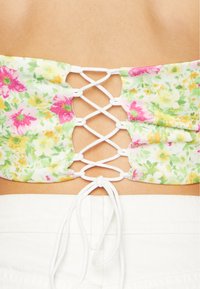 Top cropped com estampado floral em cores pastéis, apresentando um nas costas com amarração cruzada em renda e um detalhe de laço. Feito de um tecido leve.
