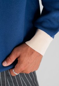 Close-up of a hand wearing a silver ring, resting near blue sweater sleeve with cream cuff and gray pinstriped pants.