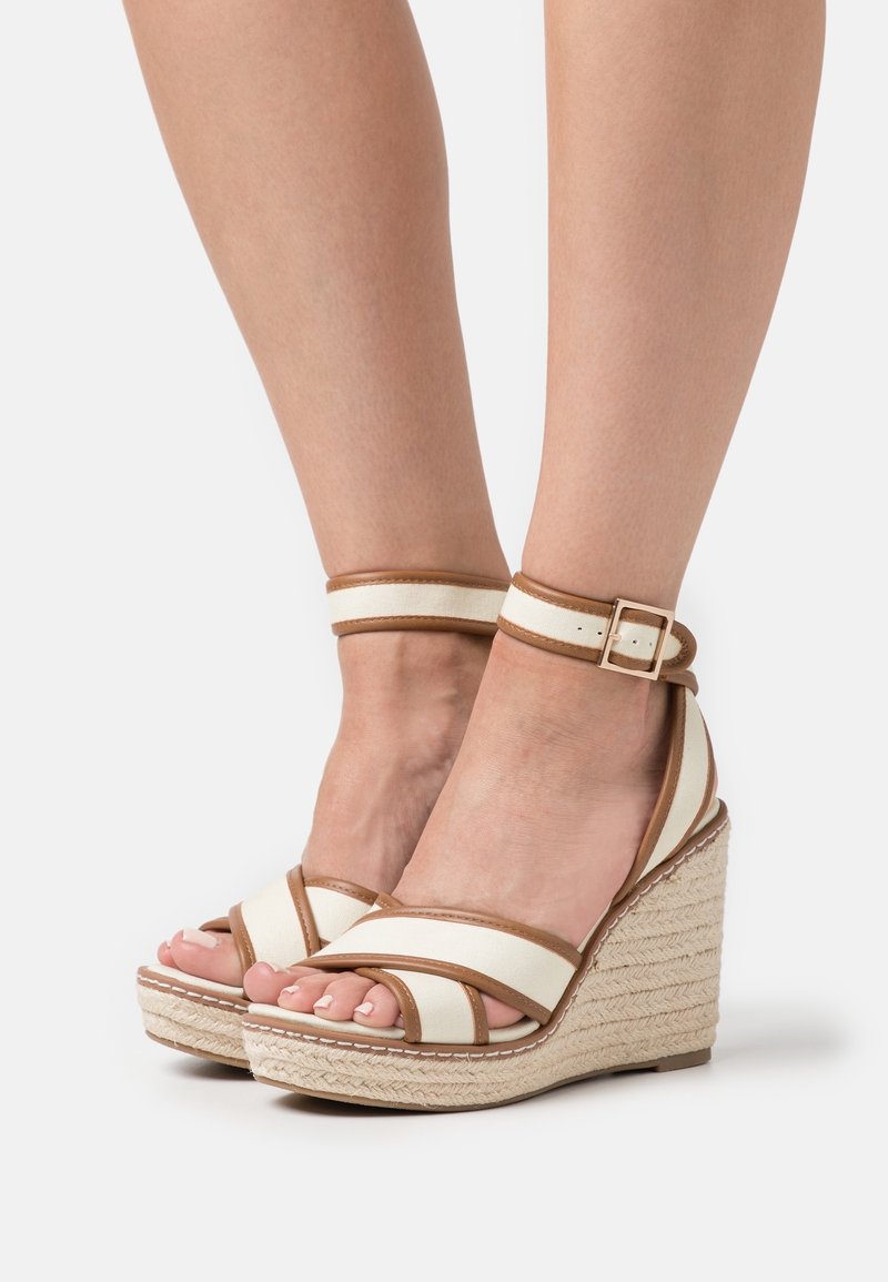 Pieds portant des sandales compensées beige et blanches avec brides autour de la cheville et semelles plateforme tressées, présentés sur un fond clair uni.