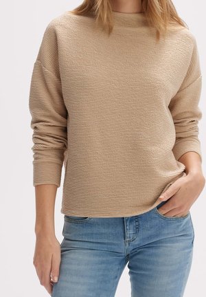 Frau trägt einen beigen, strukturierten Langarmpullover mit hellblauen Jeans, eine Hand in der Tasche, vor einem einfarbigen Hintergrund.