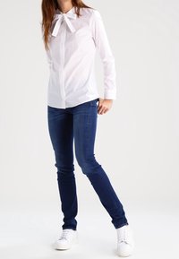 White button-up shirt with bow tie detail, paired with dark blue skinny jeans and white sneakers. Smooth fabric with a fitted design.