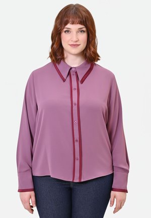 Donna con capelli rossi fino alle spalle indossa una camicetta manica lunga color malva con rifiniture rosse scure e jeans blu scuro, in piedi davanti a uno sfondo semplice.