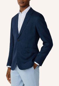 Mann trägt ein marineblaues Sakko über einem weiß gestreiften Hemd mit hellblauen Hosen und steht mit einer Hand in der Tasche.