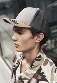 Gray cap with a beige brim, featuring a mesh back. Worn with a camouflage jacket, highlighting the structured shape and contrasting textures.