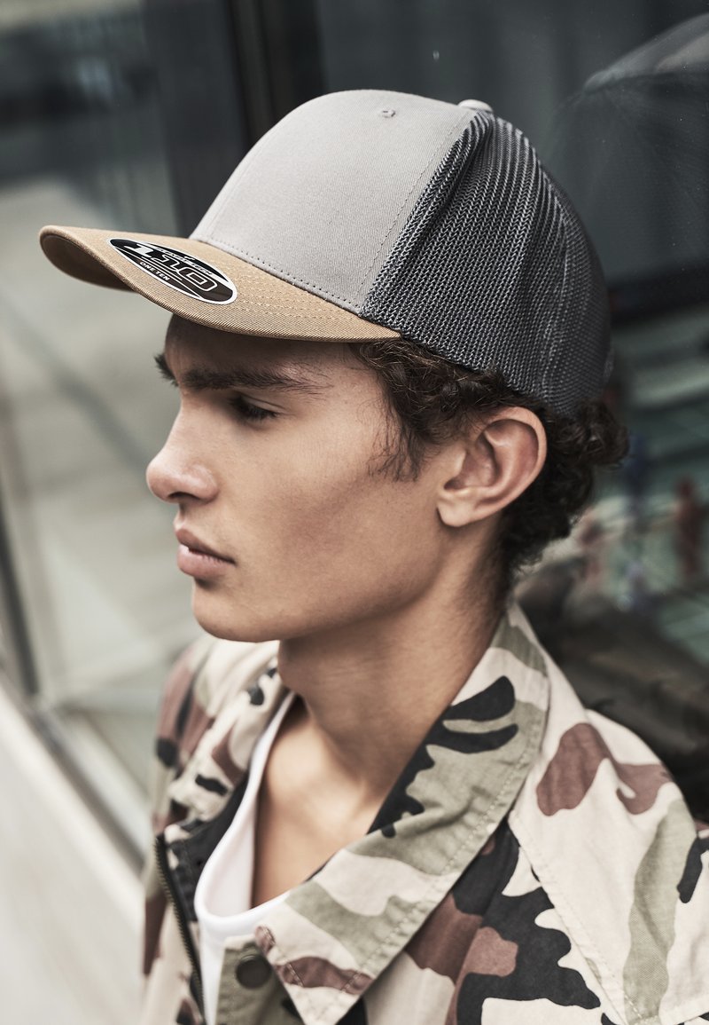 Gray cap with a beige brim, featuring a mesh back. Worn with a camouflage jacket, highlighting the structured shape and contrasting textures.