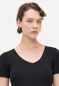 Junge Frau mit kurzen lockigen braunen Haaren und Creolen, trägt ein schwarzes V-Ausschnitt-T-Shirt vor einem schlichten weißen Hintergrund.