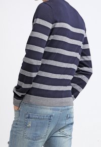 Maglione blu navy con strisce orizzontali grigie, polsini e orlo a costine, abbinato a jeans chiari a strappo, con dettaglio delle tasche.