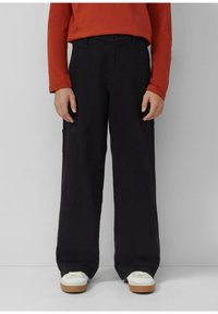 Osoba w czarnych szerokich spodniach, białych trampkach na gumowej podeszwie i czerwonej bluzie z długim rękawem, stojąca na tle jednolitego jasnego tła.