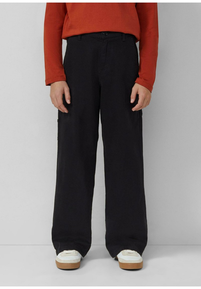 Osoba w czarnych szerokich spodniach, białych trampkach na gumowej podeszwie i czerwonej bluzie z długim rękawem, stojąca na tle jednolitego jasnego tła.