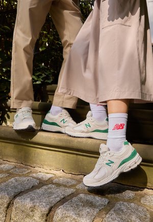 Deux personnes portant des baskets New Balance blanches et vertes et des chaussettes blanches se tiennent sur des marches en pierre à l'extérieur sous le soleil.