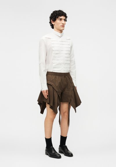 Osoba stojí čelem k předu, má na sobě bílou košili na knoflíky s horizontálními detaily a hnědé asymetrické volánkové šortky s černými botami.