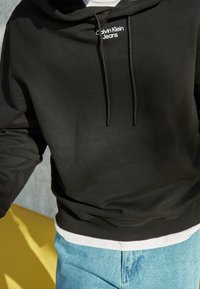 Černá mikina z měkké tkaniny s kapucí na stažení a bílým logem "Calvin Klein Jeans" na přední straně. Nositá přes bílou vrstvu.