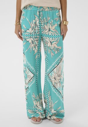 Pantalon large turquoise à imprimé floral crème, taille élastique, cordon de serrage noué, porté avec un haut beige, des bracelets dorés et des sandales beiges.