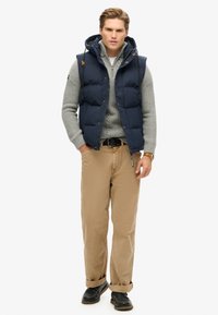 Navy puffer vest with a hoodie over a light grey knit sweater, paired with khaki pants and black shoes, featuring rolled cuffs and a belt.