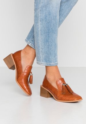 Pieds chaussés d'un jean bleu clair et de mocassins marron en cuir texturé à talon bloc avec des pampilles sur un sol blanc.