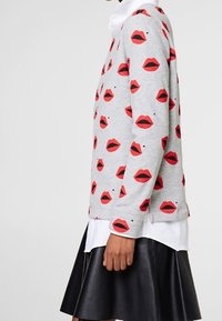 Grå sweatshirt med røde læber print, ribbede manchetter og kant, lagdelt over en hvid skjorte med krave, parret med et sort læddernederdel.