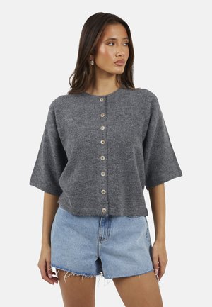Femme portant un pull gris à manches courtes avec des boutons et un short en denim bleu clair, regardant sur le côté sur un fond uni.