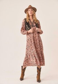 Vestido largo rosa con estampado floral, corsé negro bordado, falda con volantes y botas de vaquero marrones, acentuado por un sombrero de ala ancha.