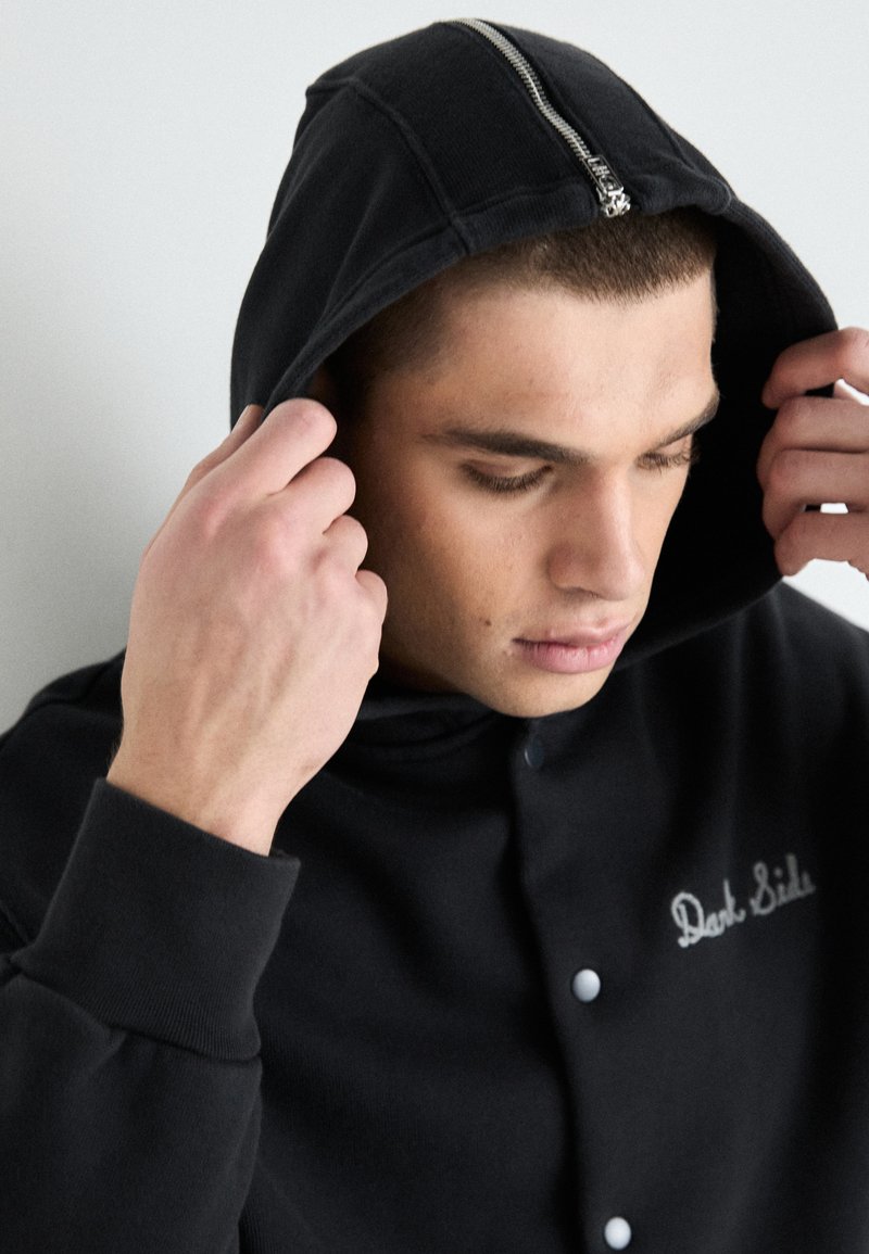 Hoodie noir avec fermeture éclair argentée, fermeture à bouton et texte brodé "Dark Side". Présente un tissu texturé et une coupe décontractée.