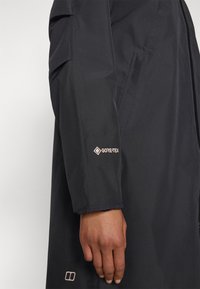 Juodas vandeniui atsparus švarkas su Gore-Tex ženklu ant rankovės. Jis pasižymi švelniu, matinio paviršiaus apdaila ir pritaikytu siluetu. Nėra matomų raštų.