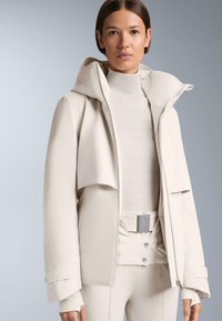 Svijetlo bež jakna s kapuljačom, ima preklop preko zatvarača, ispod se nalazi rebrasti kremasti top, a uz njega su krojene uske hlače s metalnim akcentom.