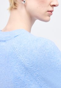 Κοντινό πλάνο προφίλ ενός ατόμου που φοράει ανοιχτό μπλε πλεκτό πουλόβερ και ασημένιο στριφτό σκουλαρίκι κρίκο σε φωτεινό φόντο.