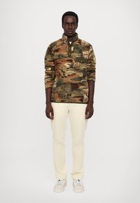 Camo-print pullover with a collar, featuring shades of green and brown, and a chest pocket. Paired with off-white pants and sneakers.