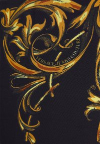 Sort stof med udsmykkede gyldne og gule barokke blomster- mønstre og teksten "VERSACE JEANS COUTURE" integreret i designet.