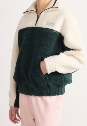 Personne portant une veste polaire vert foncé et blanc à fermeture éclair quart, avec des poches zippées, associée à un pantalon rose clair.